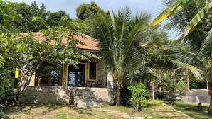 Ein Steinhaus mit Ziegeldach, einer überdachten Veranda mit Sitzgelegenheiten und einem gepflegten Garten mit Palmen und einem Kiesweg.