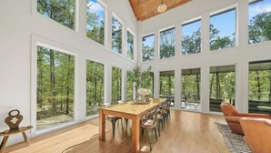 A spacious dining area with a wooden table and chairs, large windows offering a view of a forest, and a high ceiling with exposed beams.