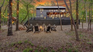 A modern house with a deck, surrounded by trees, and a fire pit with chairs.