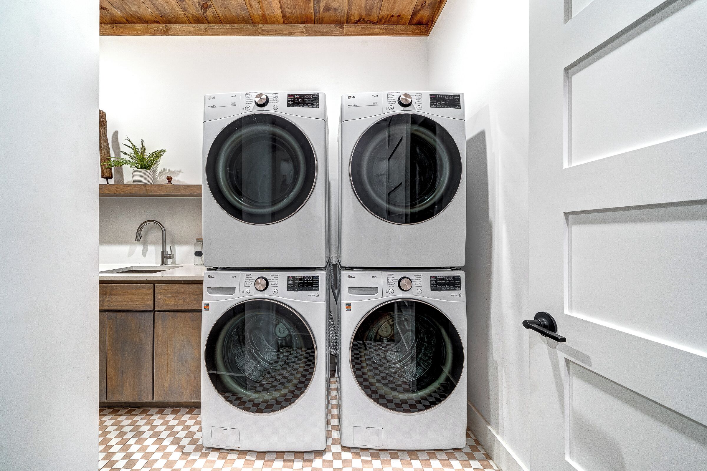 A laundry room with two stacked front-loading washing machines, a wooden shelf with a plant, a sink, and a checkered floor.