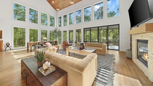 A spacious living room with a high ceiling, large windows, a fireplace, and a flat-screen TV.