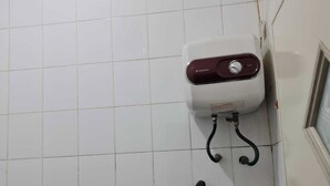 Une salle de bain avec un sol carrelé de carreaux blancs, des toilettes blanches dotées d’un panneau de commande rouge, une douchette noire et un seau bleu.