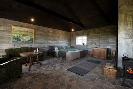 A rustic room with a stone floor, a bed with a green blanket, a wooden table with candles, a sofa, a painting on the wall, and a fireplace.