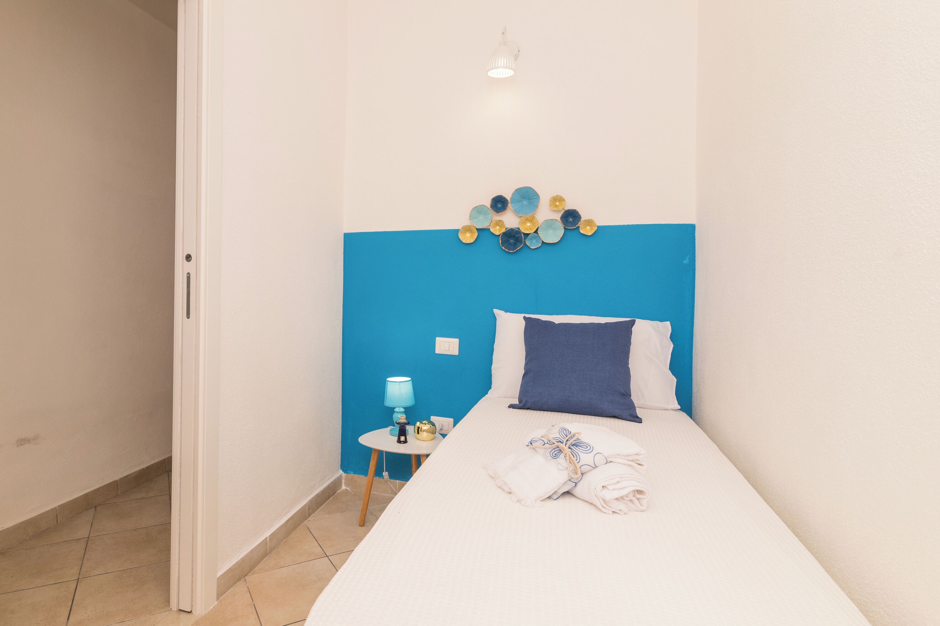 A hotel room with a bed, a blue headboard, a small bedside table with a lamp, and a wall decoration.