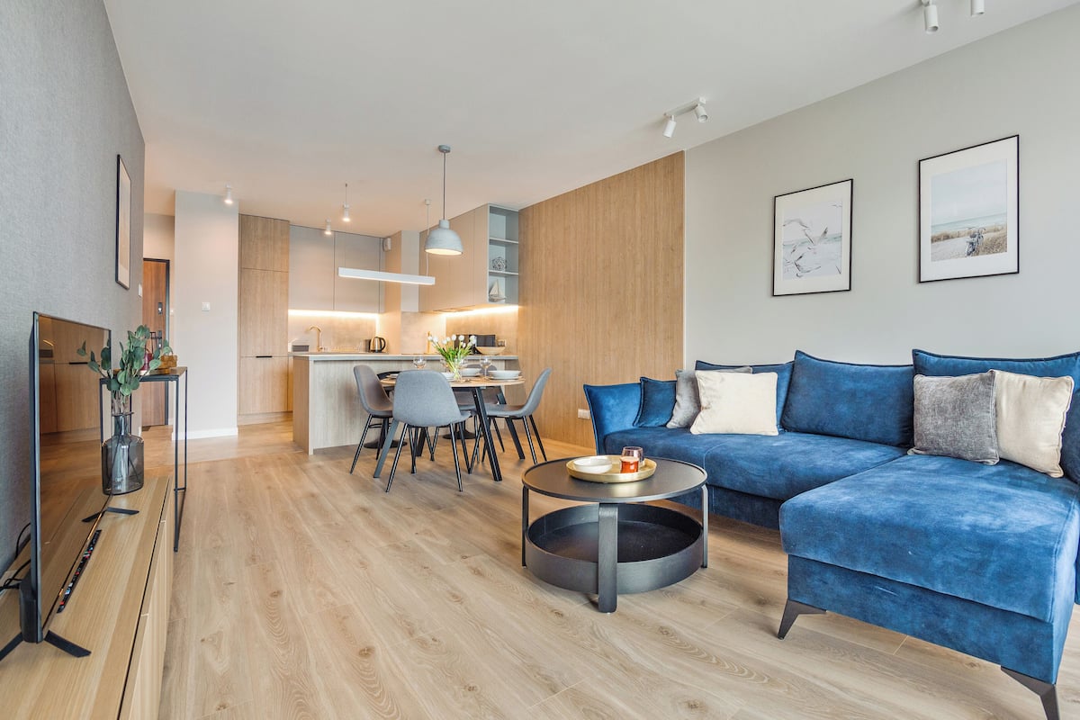 A modern living room with a blue sofa, a dining area with a table and chairs, and a kitchen in the background.