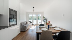 A modern kitchen with built-in appliances, a dining area with a wooden table and chairs, and a view of a green building through large windows. - Apartamenty Sun&Snow Baltea (Gdansk)