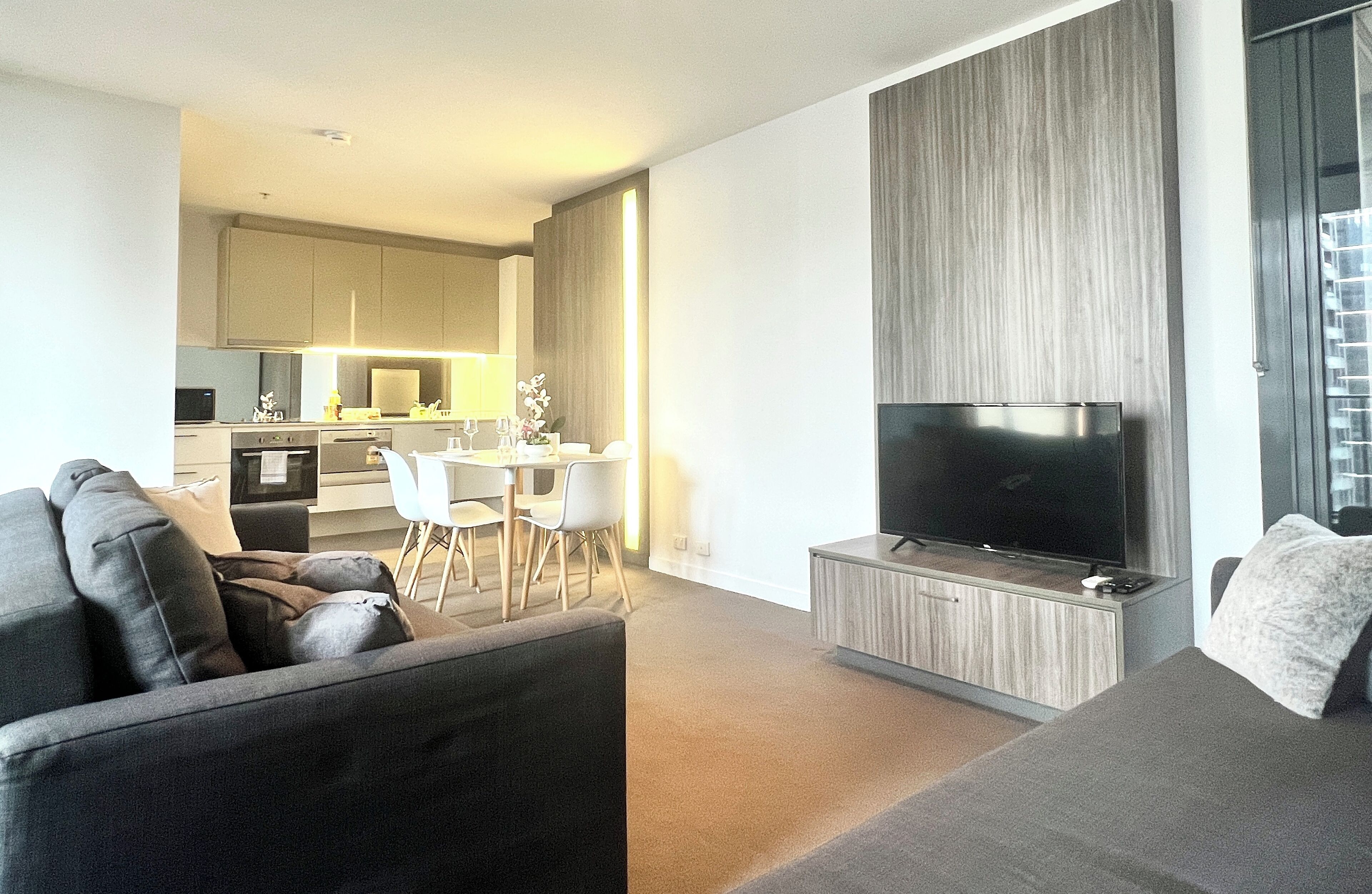 A modern living room with a grey sofa, a flat-screen TV, and a dining area with white chairs and a table.