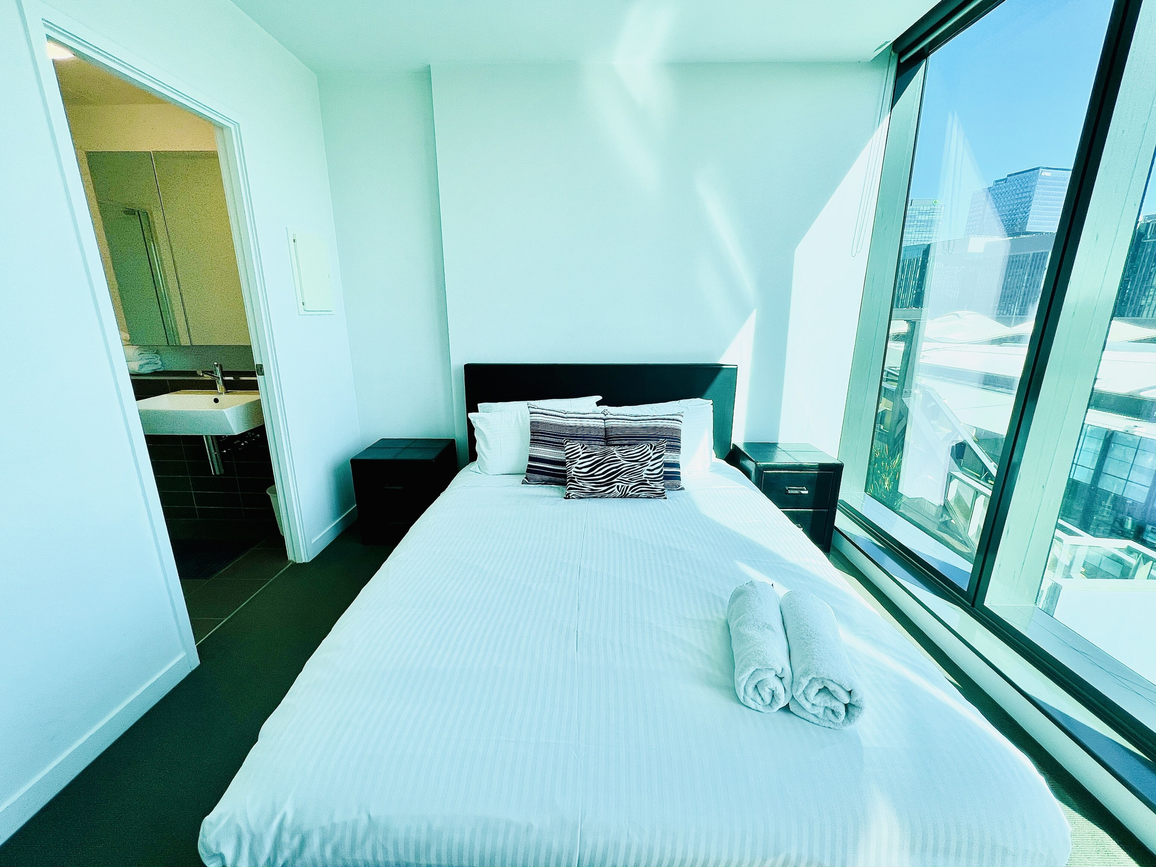 Une chambre d’hôtel avec un grand lit, une table de chevet, une petite table, une salle de bain avec un lavabo et une vue sur la ville par une grande fenêtre.