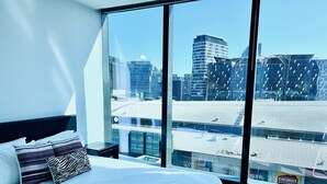 Une chambre d’hôtel moderne dotée d’un grand lit, d’une table de chevet et offrant une vue sur le paysage urbain grâce à de grandes fenêtres du sol au plafond.