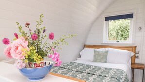 A bedroom with a wooden bed, a green patterned bedspread, and a blue bowl with flowers on a white bedside table. - Ash (Saltash)