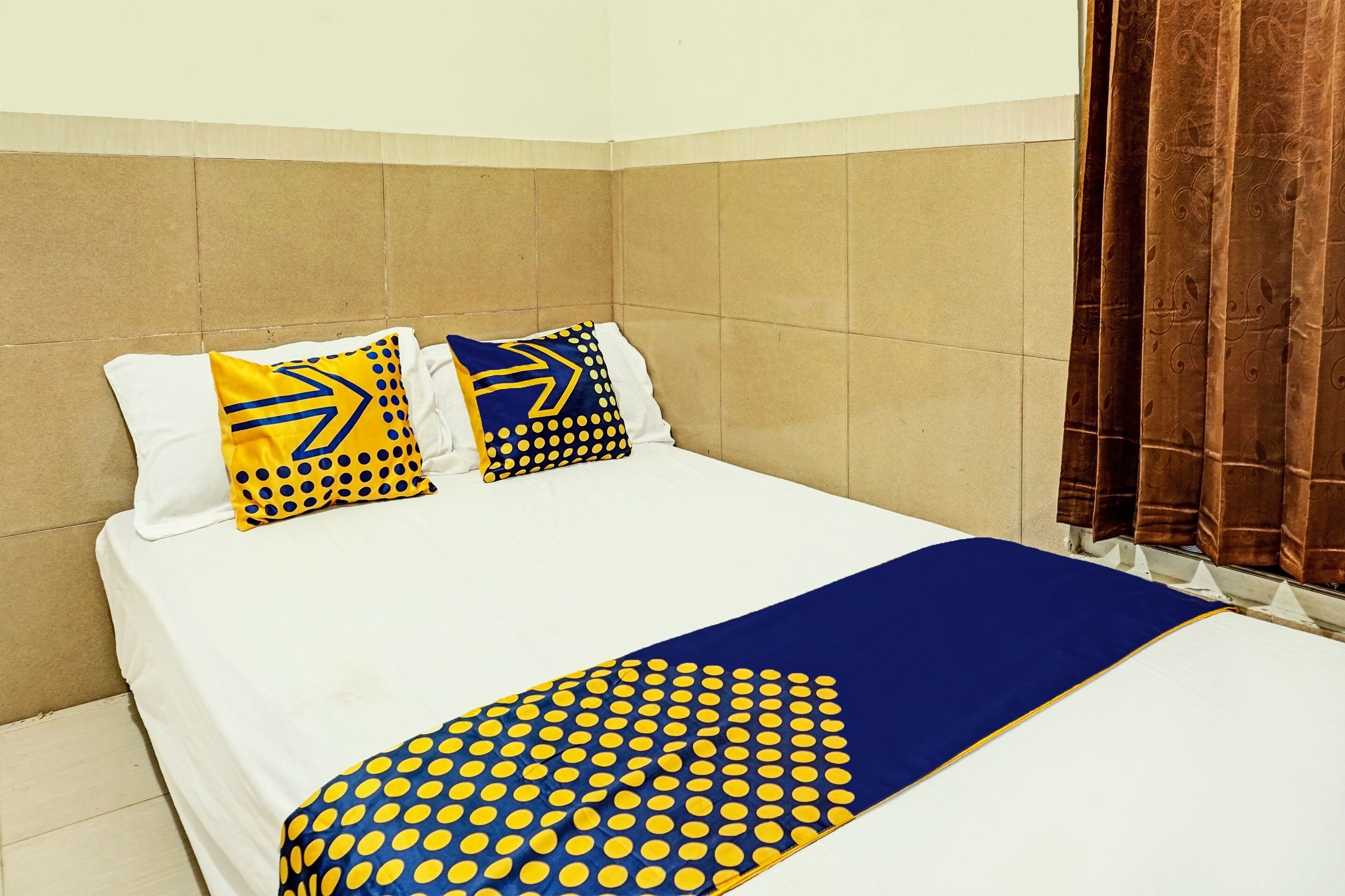 A neatly made bed with a white and blue polka dot bedspread, two matching pillows, and a brown curtain on the window.