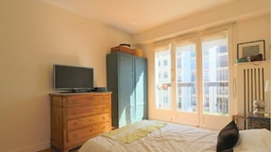 Een slaapkamer met een bed, een dressoir, een televisie erboven en een raam met uitzicht op gebouwen.