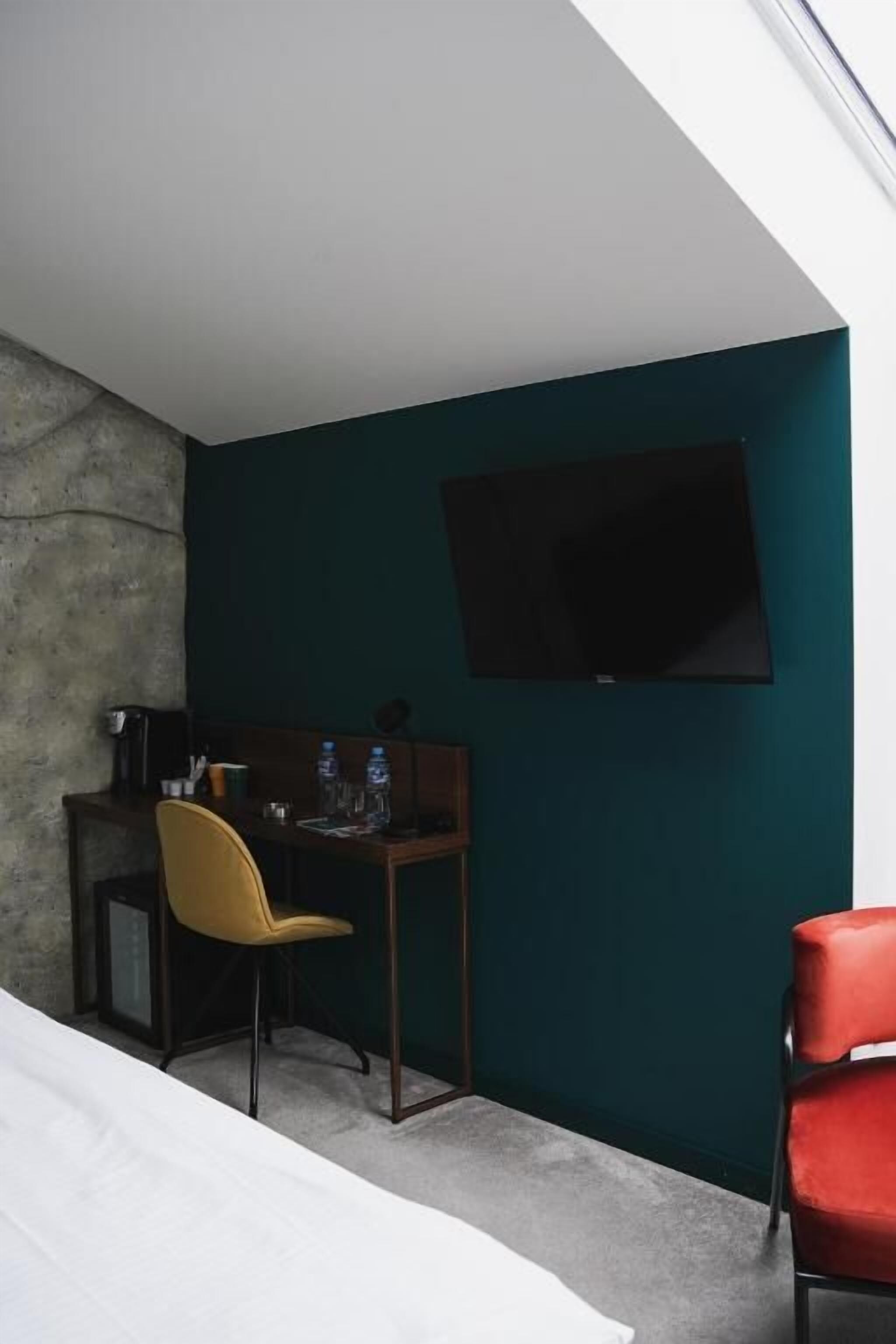 A compact hotel room with a desk, a yellow chair, a red chair, a bed, and a TV mounted on the wall.
