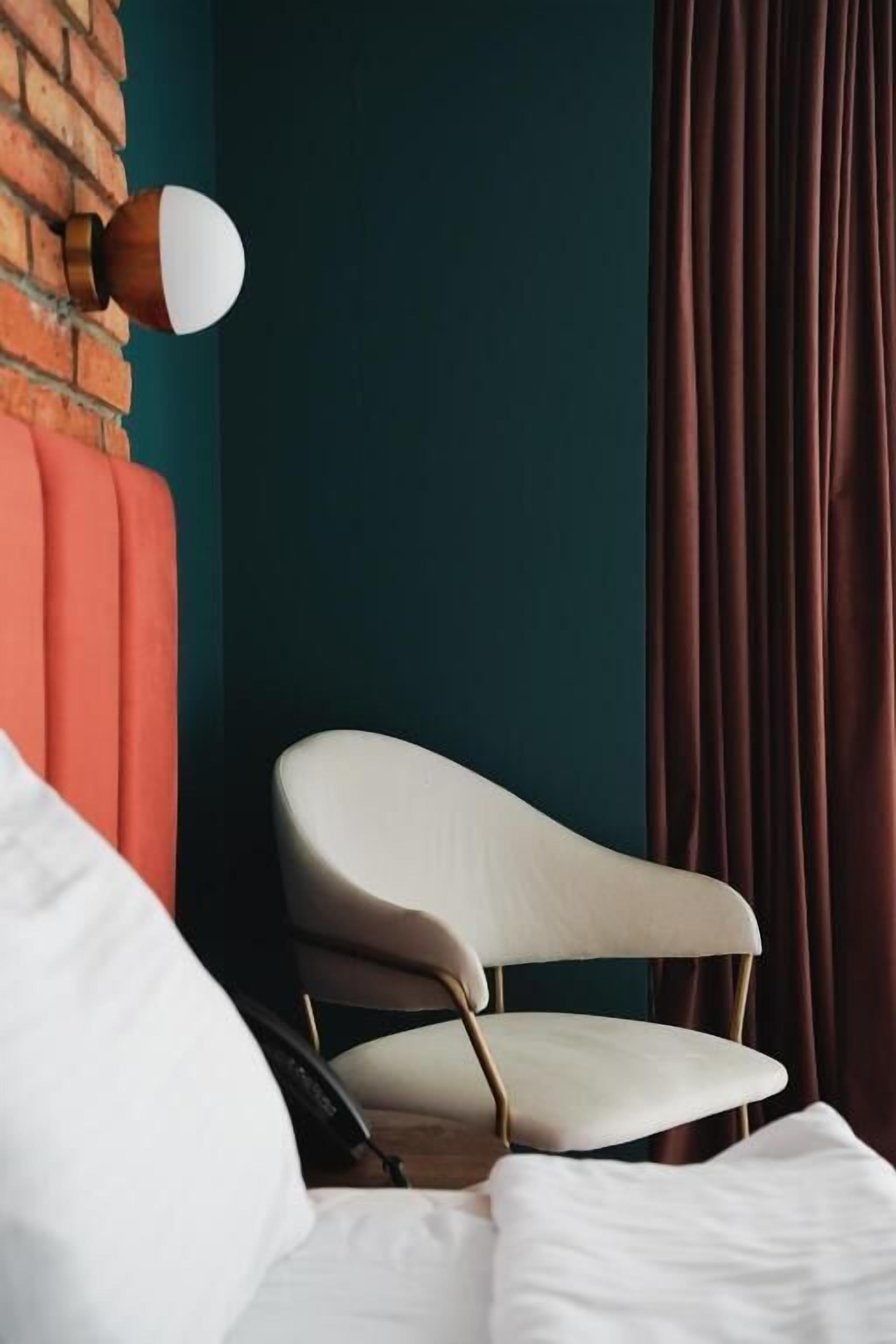 Une chambre d’hôtel moderne avec un mur d’accent en briques, un fauteuil blanc recouvert de tissu, un lit avec une literie blanche et un mur vert foncé.