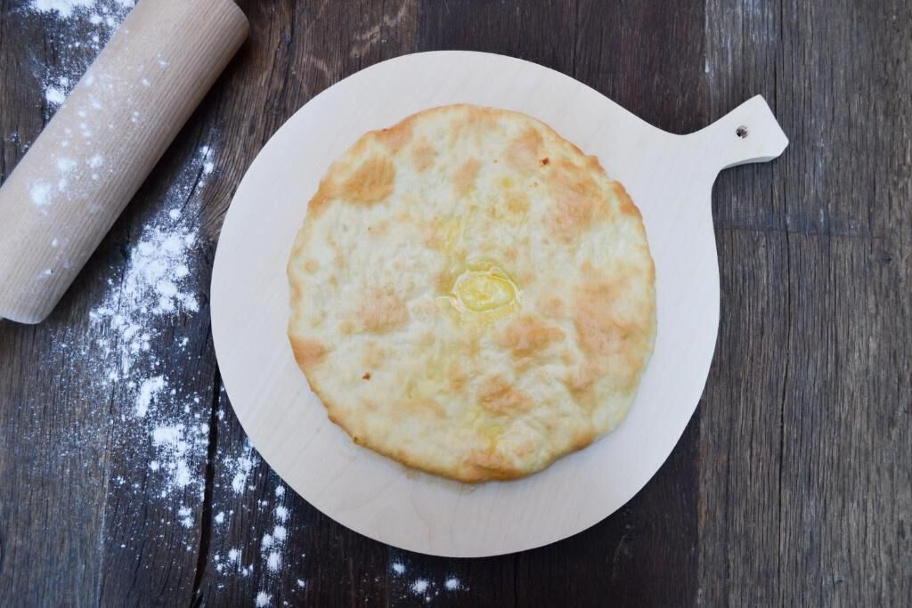 A round, flat, golden-brown flatbread on a white plate with a handle, placed on a wooden surface with flour scattered around.