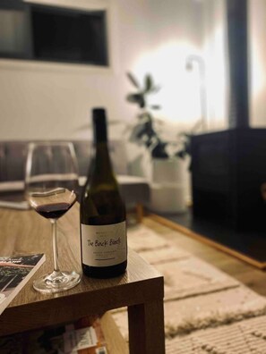 A bottle of wine and a glass on a wooden table in a living room with a white couch and a plant in the background.