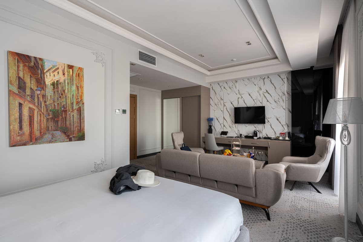 A hotel room with a bed, a sofa, a desk, a TV, and a painting on the wall.
