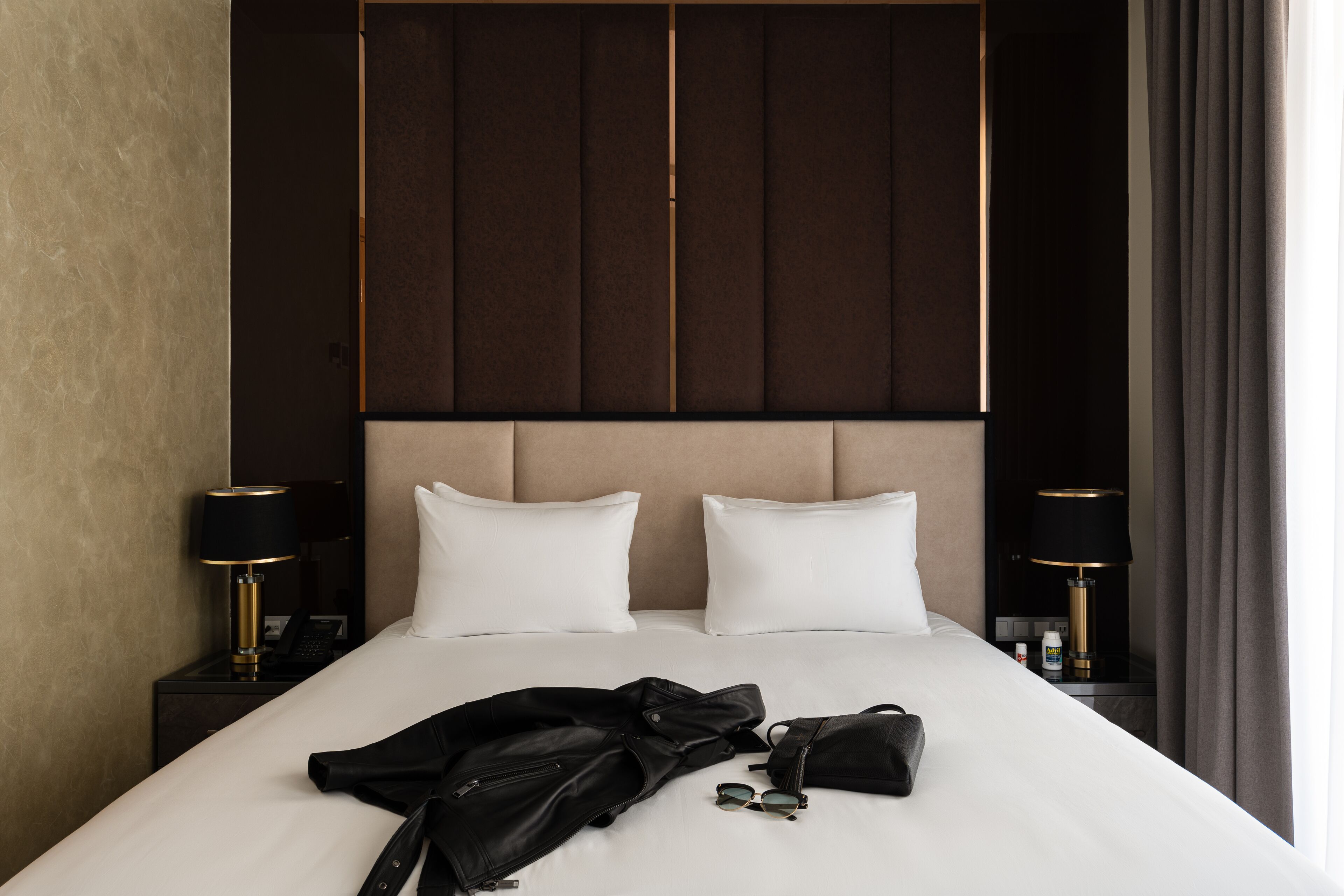 A neatly made bed with white linens, two pillows, a black jacket, and a black bag on the bed. There are two bedside tables with lamps and a small bottle on one of them.