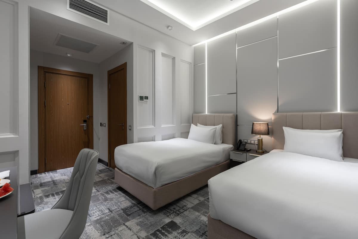 A hotel room with two beds, a desk with a chair, and a bedside table with a lamp.