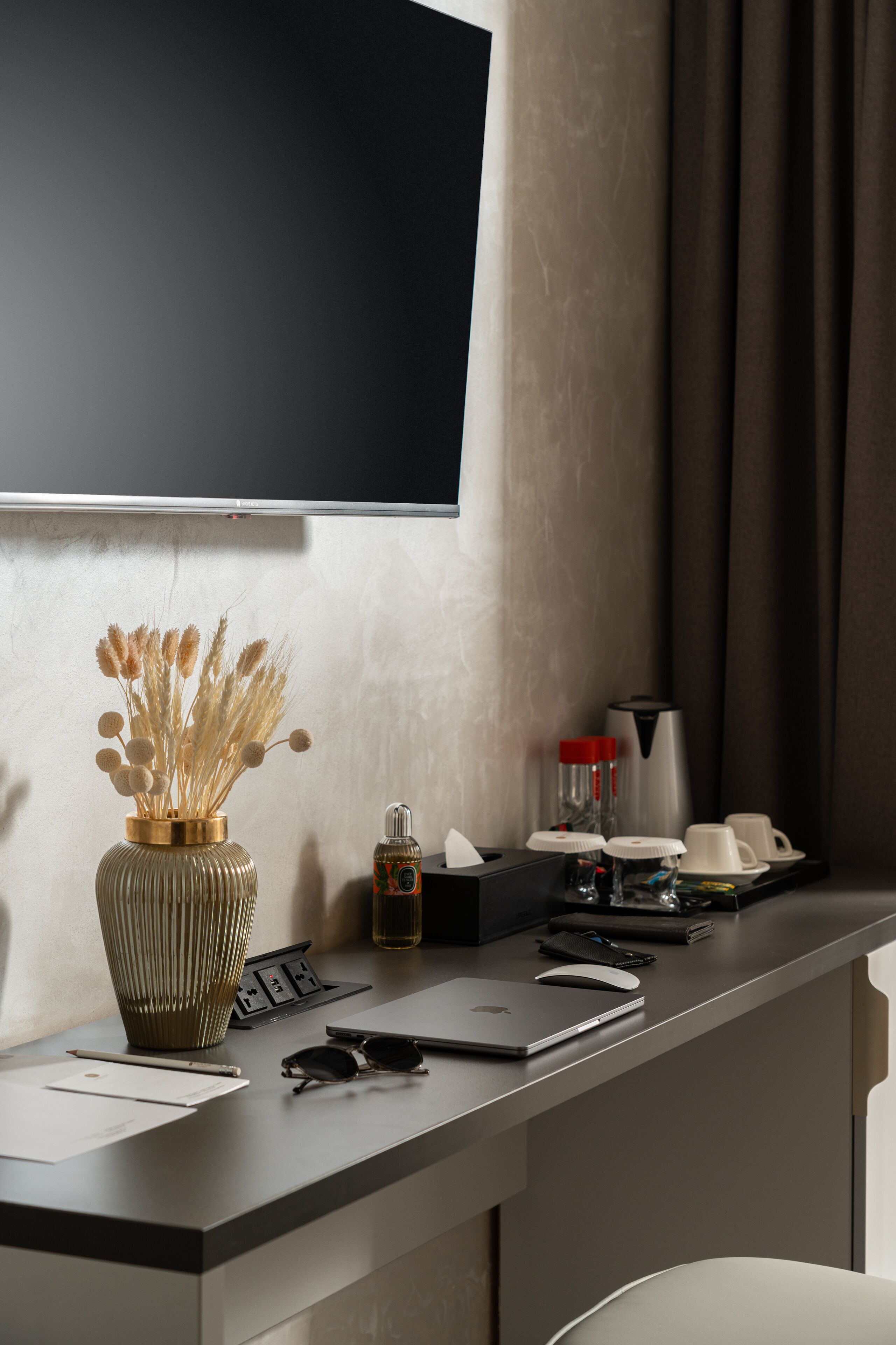 A modern hotel room with a flat-screen TV mounted on the wall, a desk with a laptop, a vase with dried flowers, a bottle of hand sanitizer, and a coffee maker.