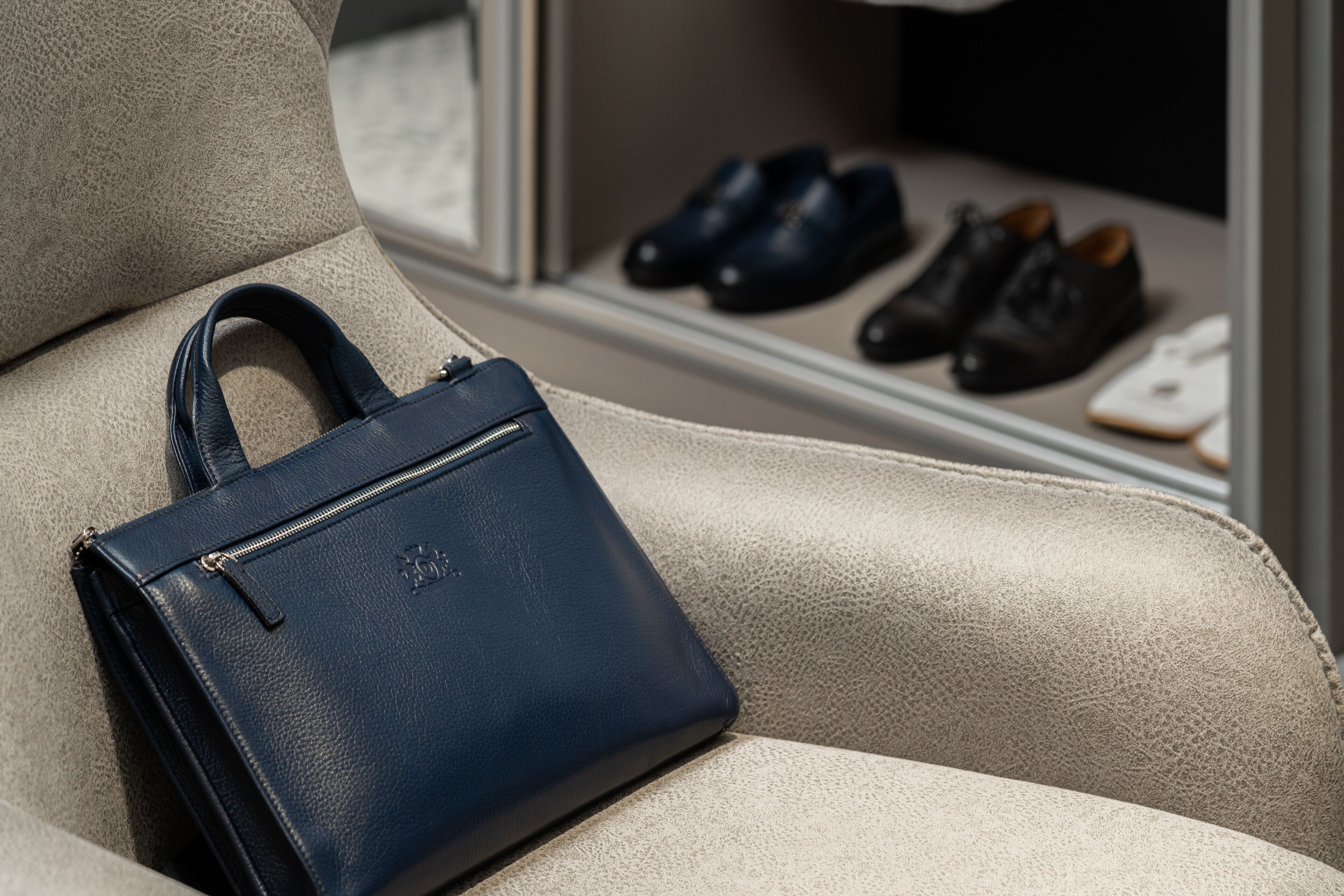 Un bolso azul oscuro sobre un sofá de cuero color beige, con un par de zapatos negros y un objeto blanco al fondo.