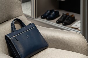 Un bolso azul oscuro sobre un sofá de cuero color beige, con un par de zapatos negros y un objeto blanco al fondo.