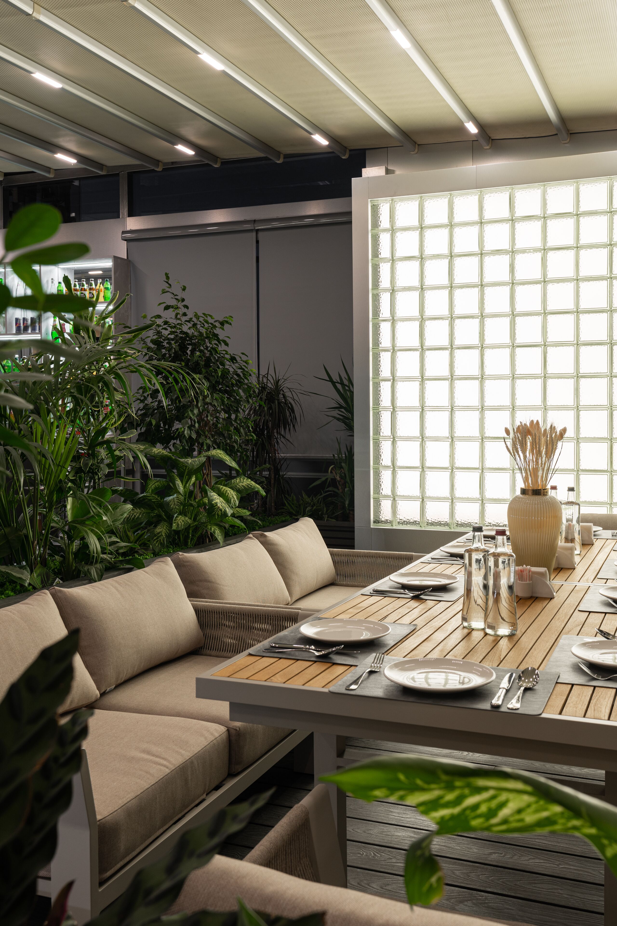 A dining area with a wooden table set for two, surrounded by a sofa and potted plants.