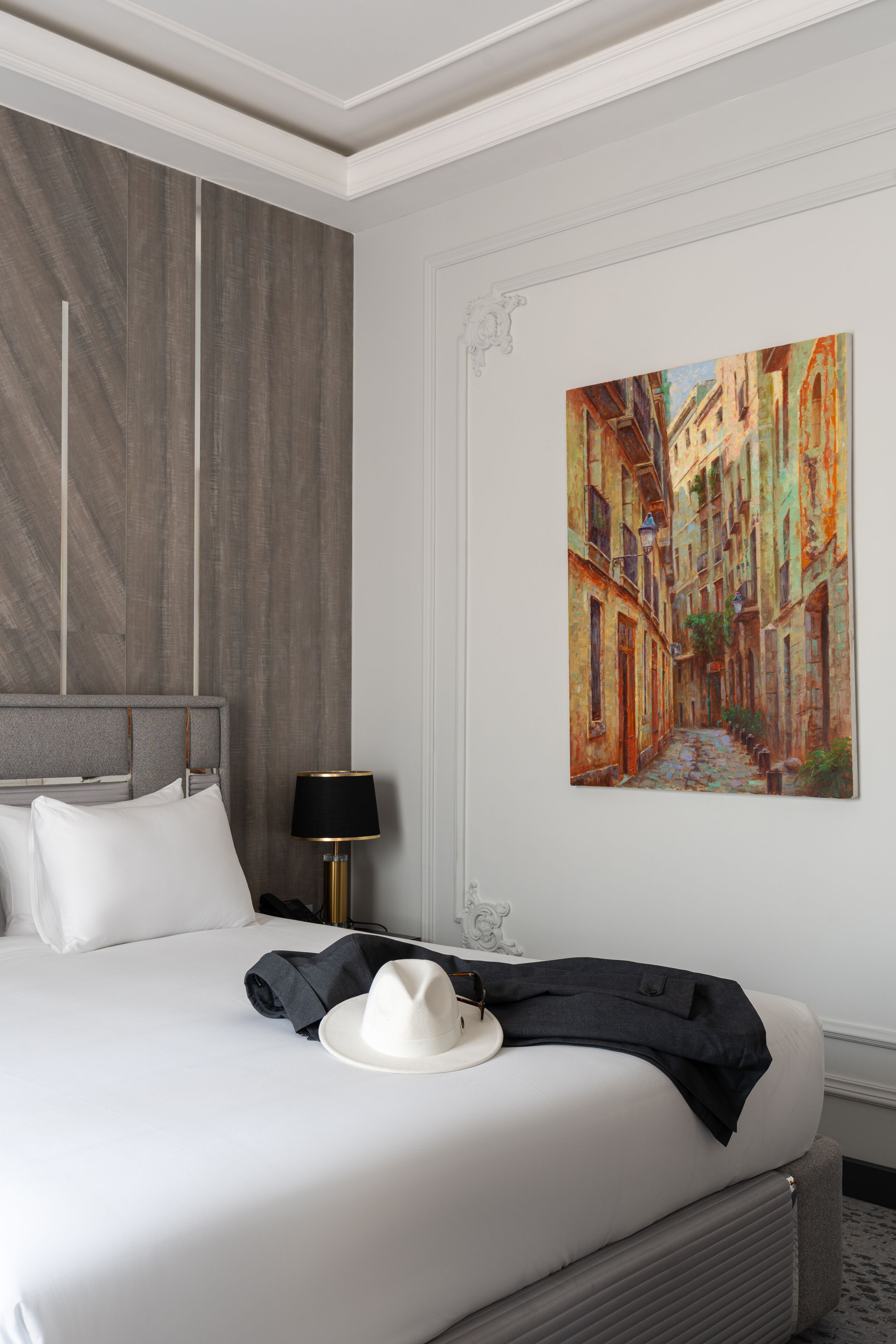Un cuarto de hotel con una cama bien hecha, un cuadro de una escena callejera en la pared y una lámpara junto a la cama.