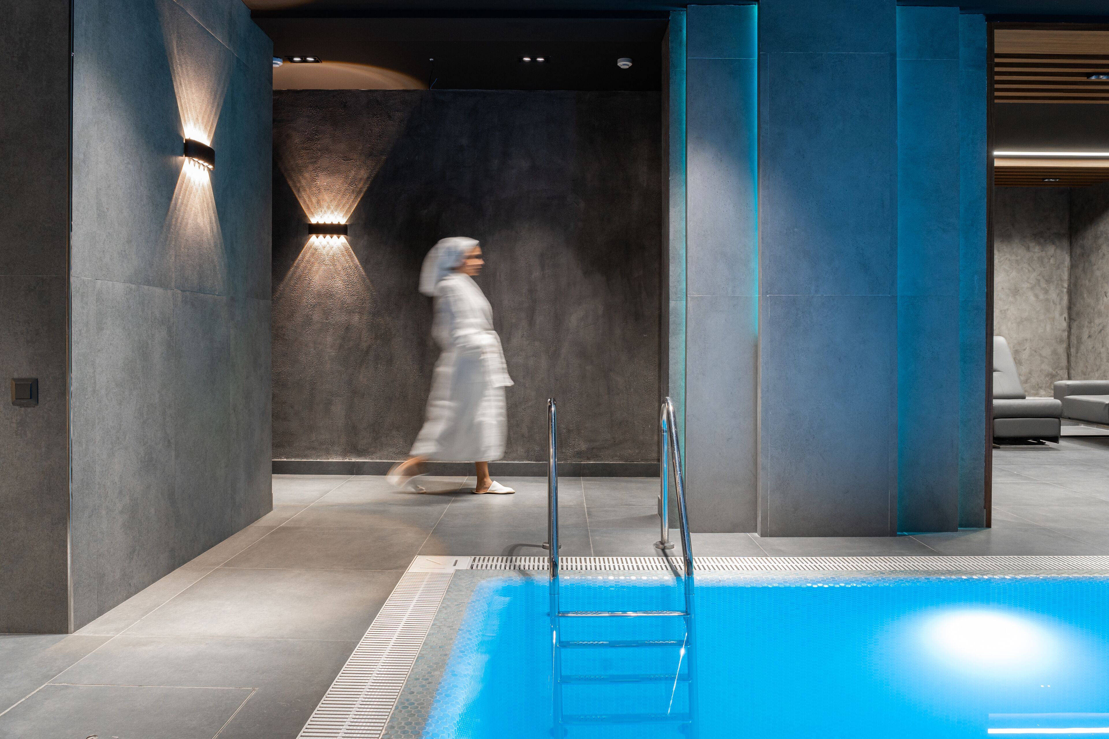 A modern indoor pool area with a clear pool, a ladder, and a person walking in the background.