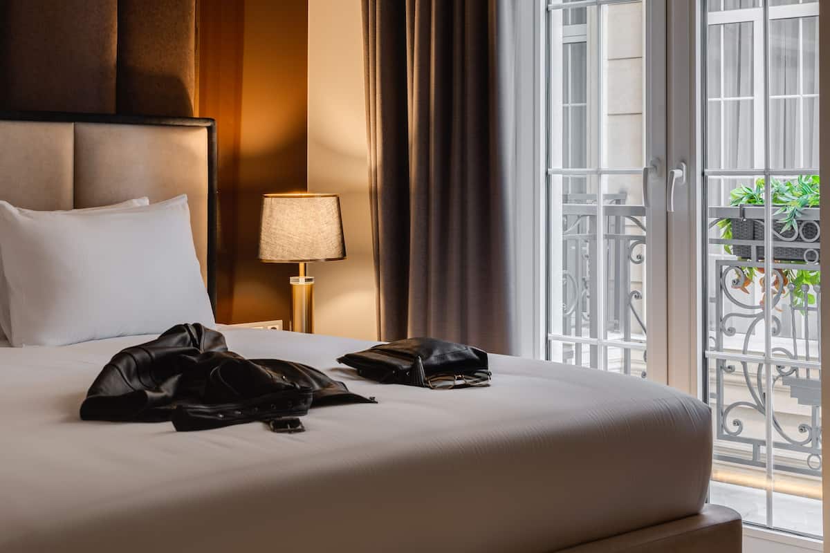 A hotel room with a large bed, a nightstand, a lamp, and a view of the outside balcony.