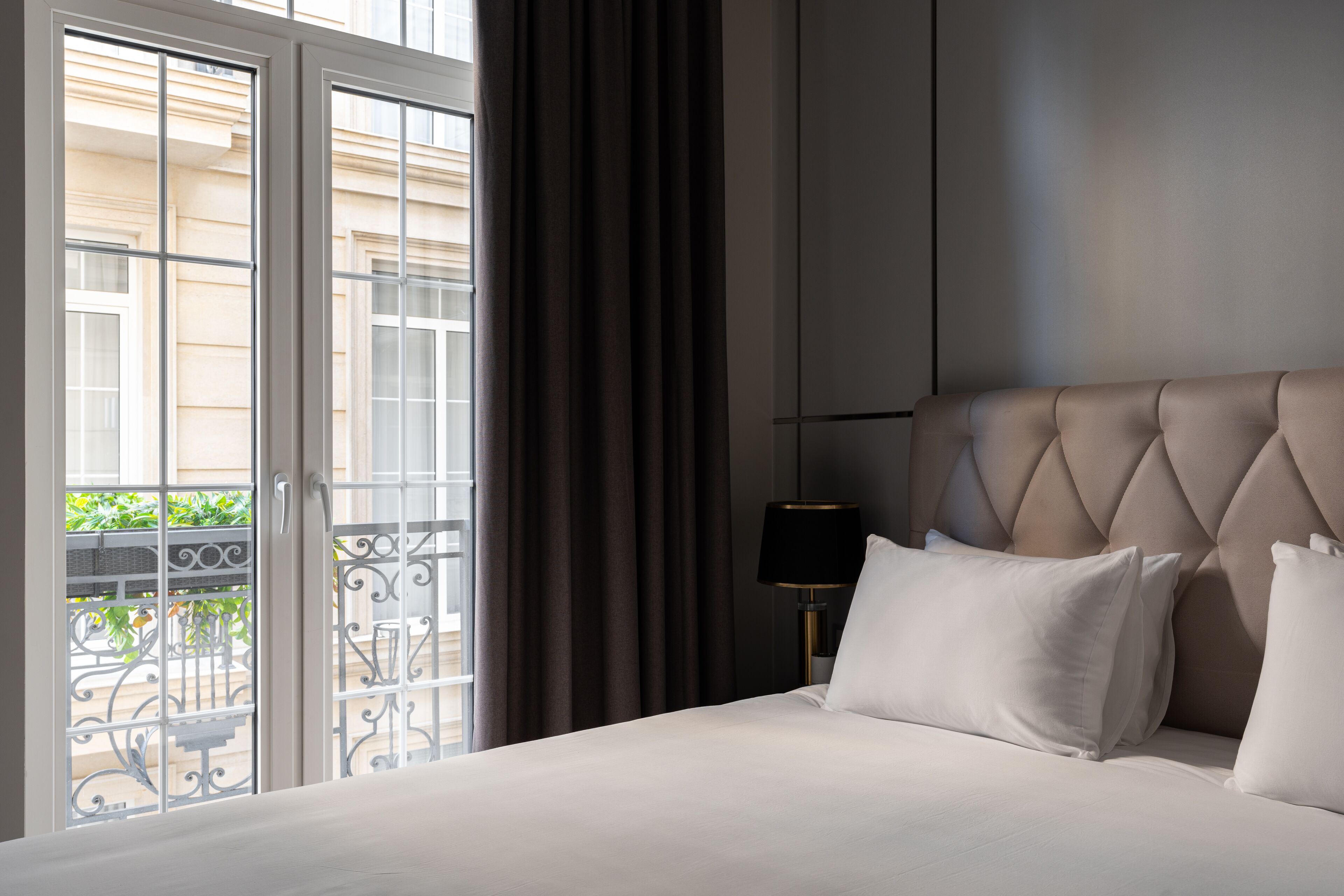 A hotel room with a bed, a nightstand with a lamp, a view of a balcony with a railing, and a window with curtains.