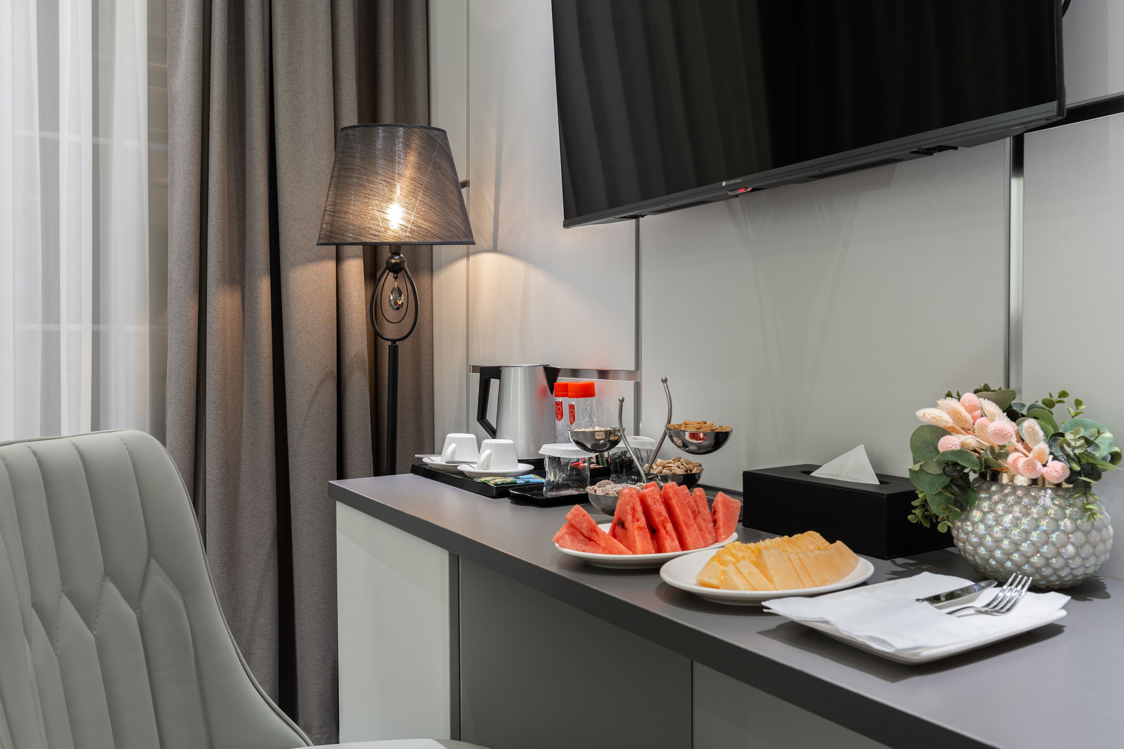 A hotel room with a desk, a flat-screen TV, a lamp, a vase with flowers, and a plate of food.