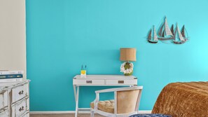 Una habitación con una pared verde azulado, un escritorio blanco con cajones, una lámpara y una cama con una manta marrón.