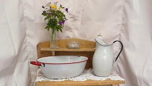 Ett rustikt träskåp med en vit skål och en vit kanna ovanpå, en liten vas med blommor och ett vitt bord med en vit duk.