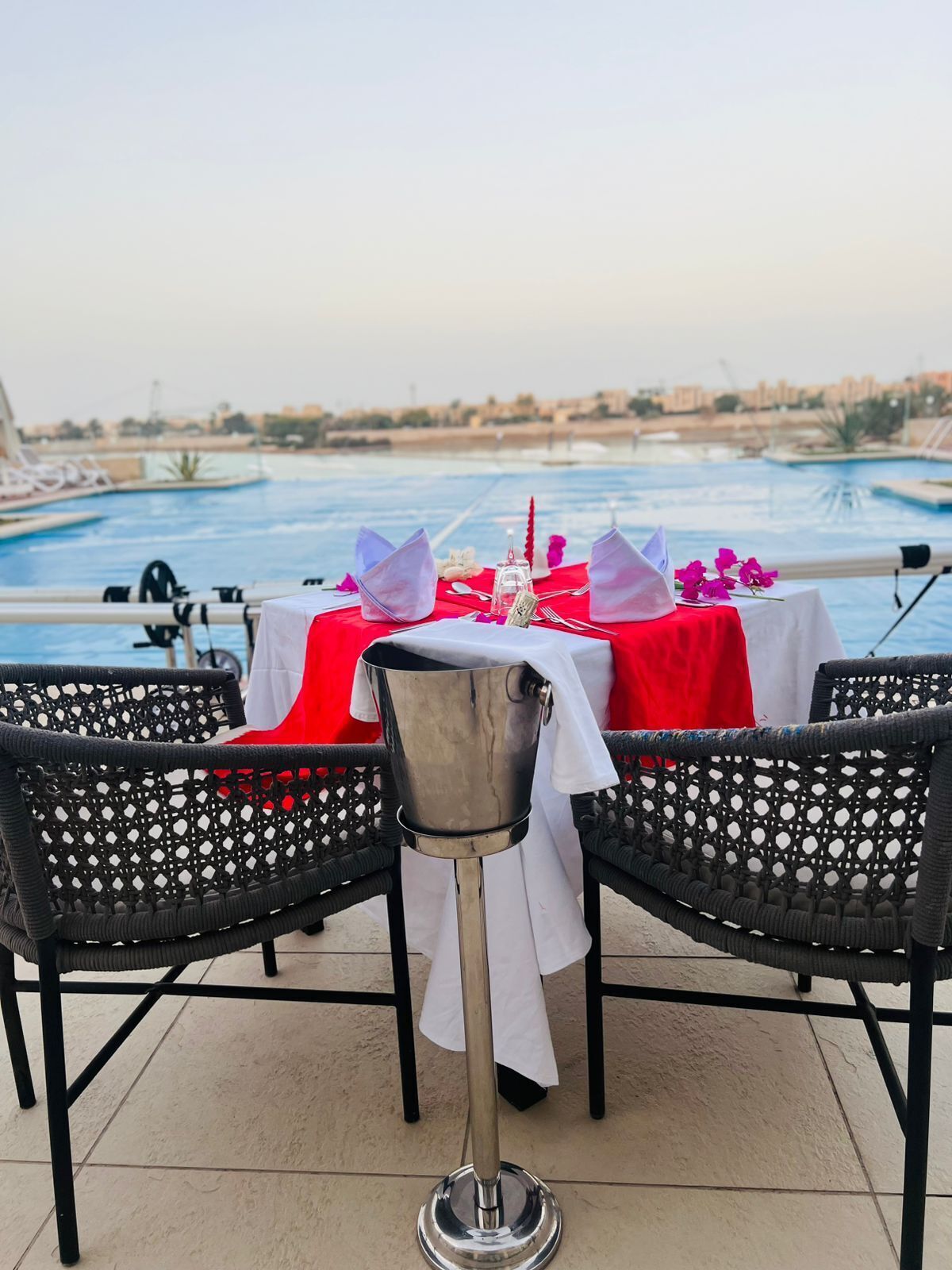 A dining table set for two with a view of a swimming pool and distant buildings.