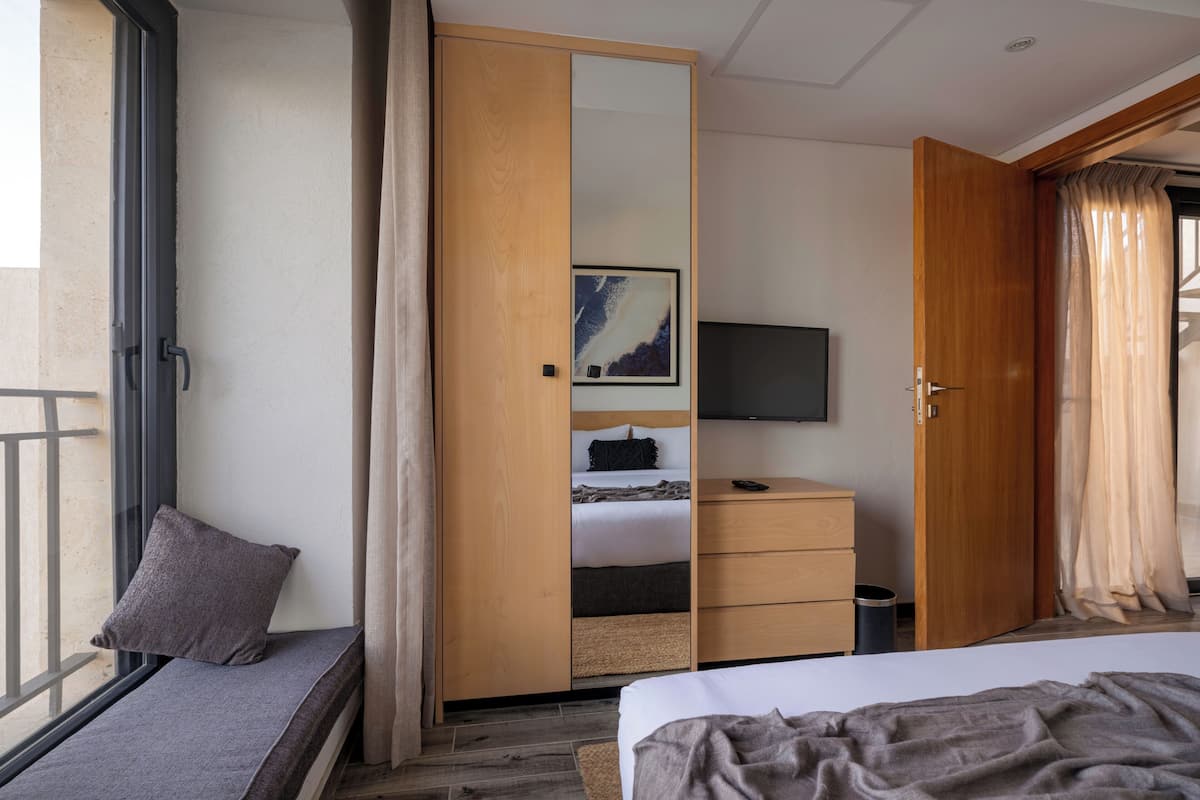 Hotellihuone, jossa on sänky, televisio, puinen vaatekaappi, yöpöytä, roskakori ja ikkuna, josta on näkymä.