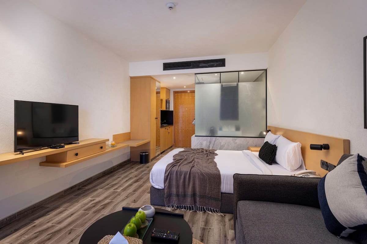 Moderni hotellihuone, jossa on taulutelevisio, sohva, sängynpäätyyn varustettu sänky, yöpöytä ja suuri peili.