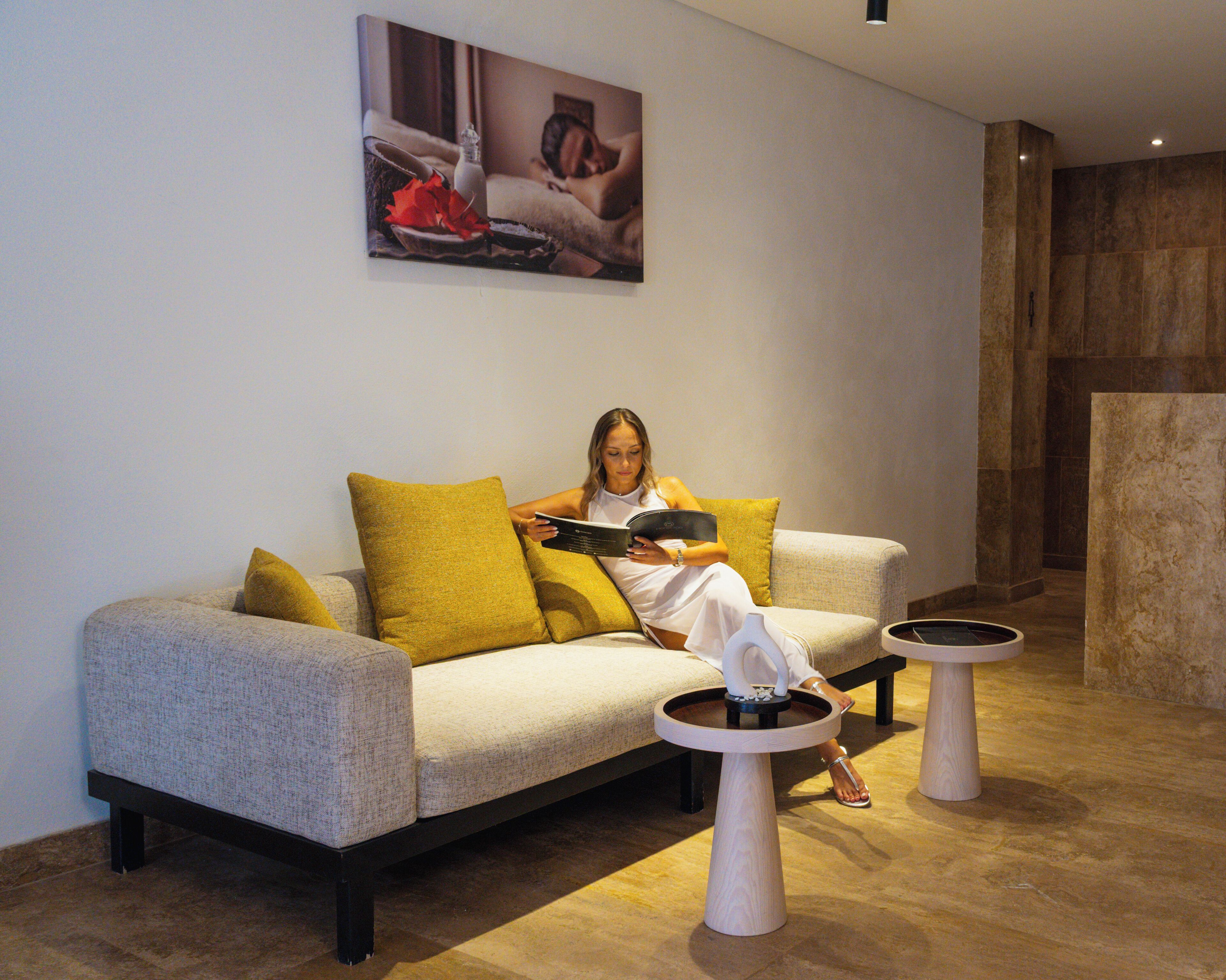 A woman sitting on a sofa with a book, surrounded by two small tables and a painting on the wall.