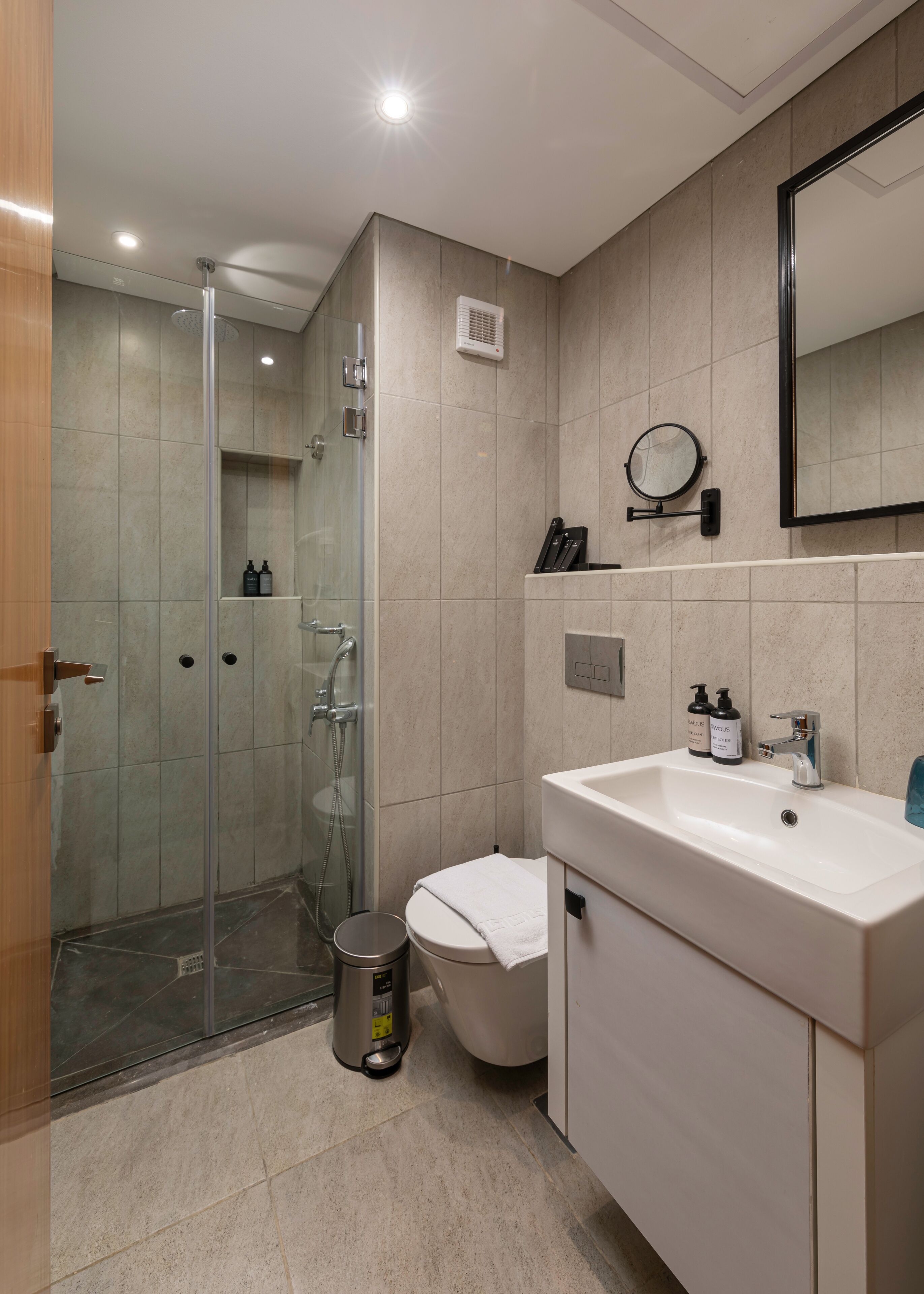 A modern bathroom with a glass shower enclosure, a toilet, a sink with a mirror, and a trash bin.