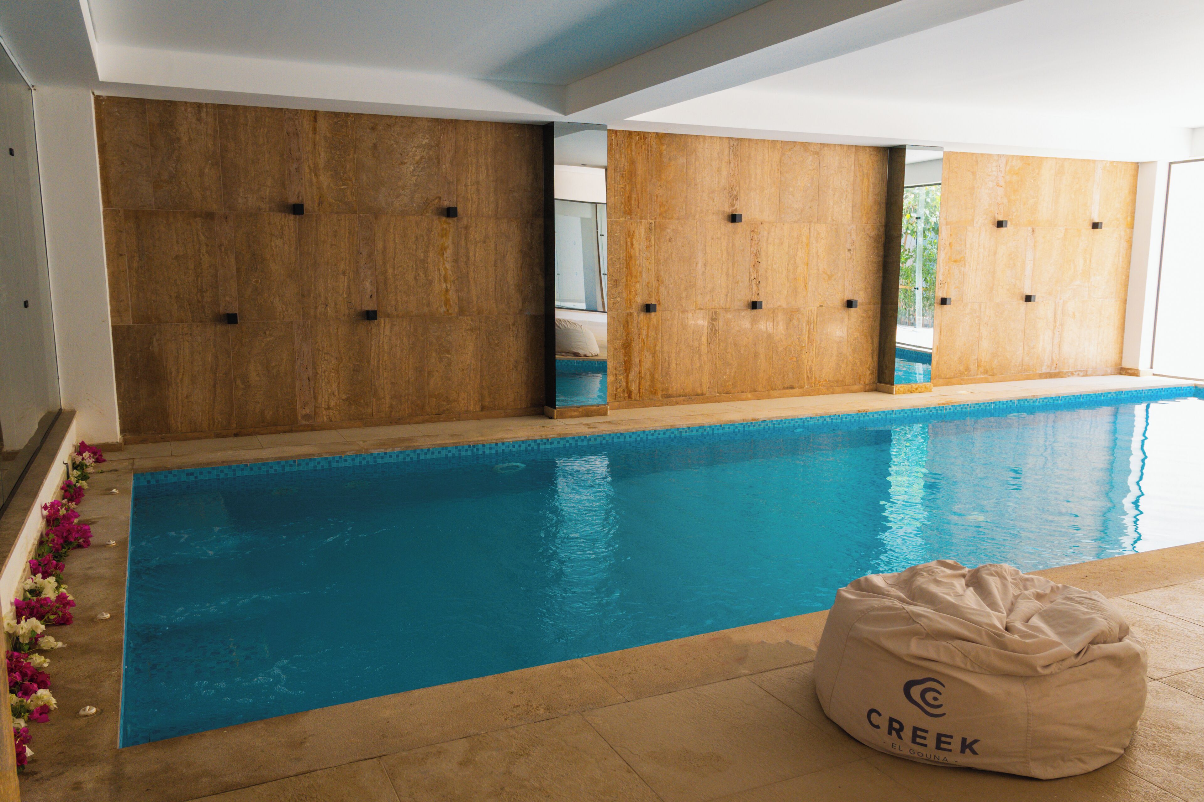 A swimming pool with wooden paneling on the walls, a bean bag chair labeled "CREEK EL BOUJA" in the foreground, and a tiled floor.