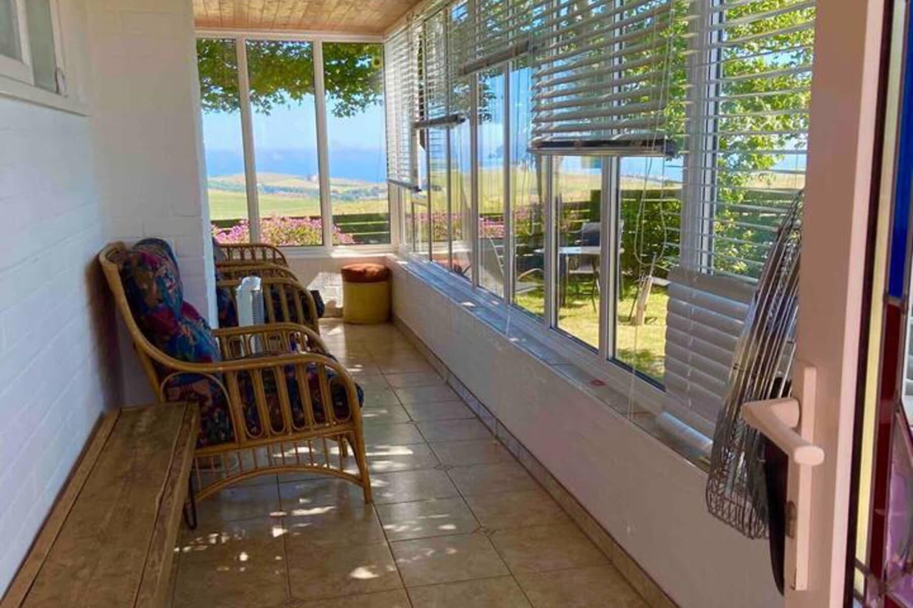 Une pièce baignée de lumière naturelle, avec vue sur l’extérieur, comprenant un fauteuil en osier, une petite table et une fenêtre équipée de stores.