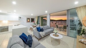 A modern living room with a grey sofa, a coffee table, and a view of a sunset through large glass doors. - Epitome of luxury - absolute waterfront penthouse (Cannonvale)