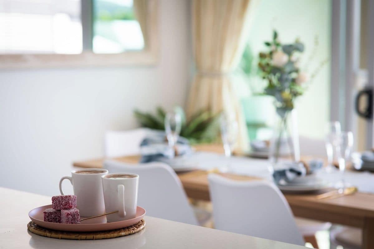 Ein Esstisch mit einer Speisenplatte und zwei Tassen, vermutlich Kaffee, in einem hellen Raum mit Fenster und einer Pflanze im Hintergrund.