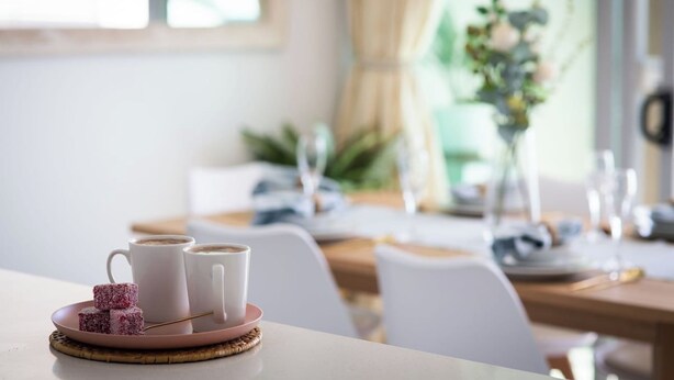 Una mesa de comedor con un plato de comida y dos tazas, probablemente de café, en una habitación luminosa con una ventana y una planta al fondo.