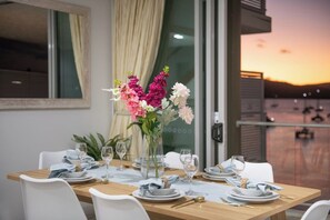 A dining table set for two with a vase of flowers, white chairs, and a view of the sunset.