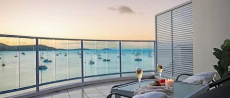 A balcony with a glass table, two lounge chairs, a candle, and a glass of champagne.