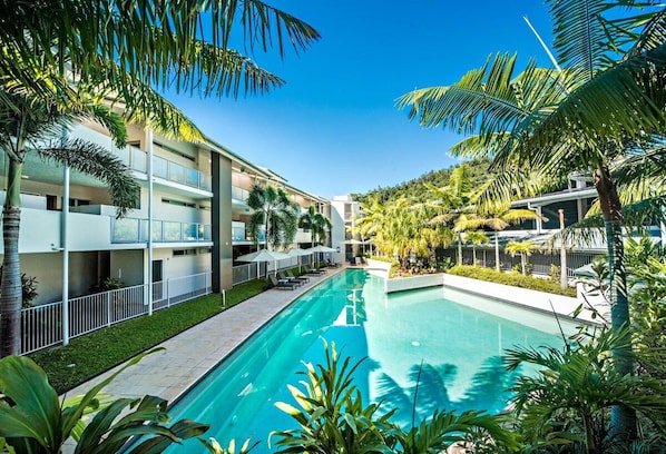 A hotel with a swimming pool surrounded by palm trees and a hilly landscape.