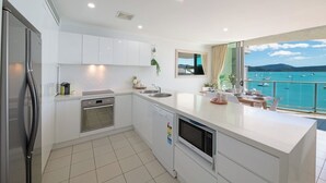 A modern kitchen with white cabinets, a built-in oven, and a dishwasher. There is a large window with a view of a body of water and boats. - Epitome of luxury - absolute waterfront penthouse (Cannonvale)