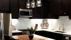 Moderni keittiö, jossa on tummat puukalusteet, ruostumattomasta teräksestä valmistetut kodinkoneet, puinen keittiösaareke baarituoleineen, riippuvalaisimet ja ruukussa oleva kasvi.