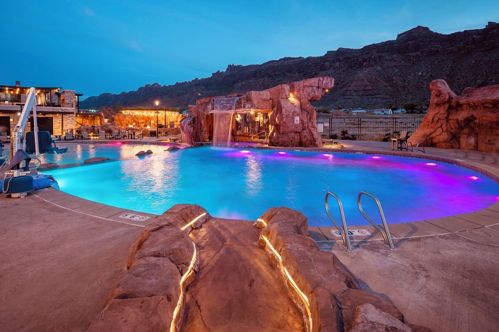 A swimming pool with a waterfall feature, surrounded by rock formations and a building with outdoor seating.